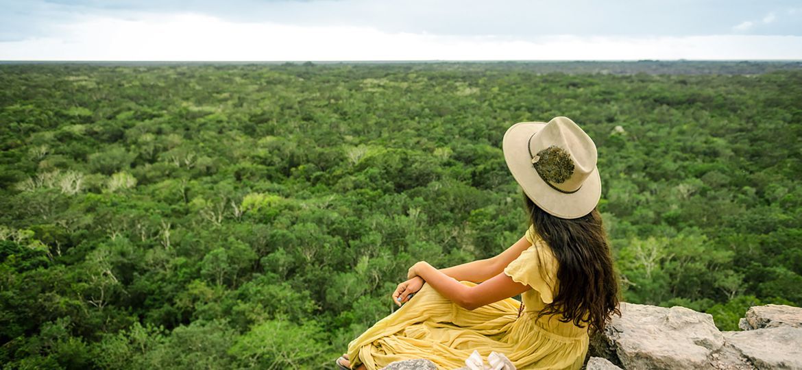 Mujer joven con vestido amarillo viendo al horizonte de la jungla maya desde la cima de la piramide de coba