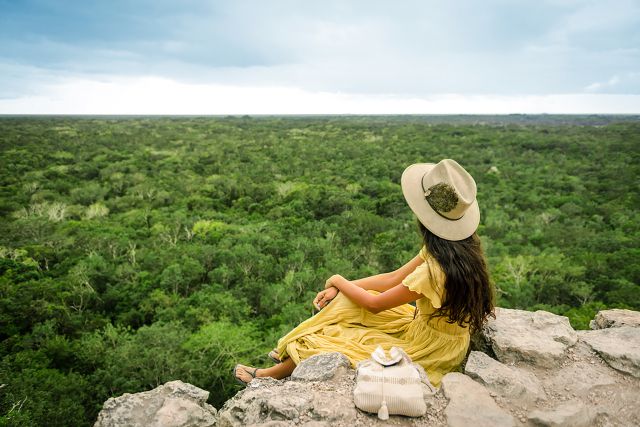 Mujer joven con vestido amarillo viendo al horizonte de la jungla maya desde la cima de la piramide de coba